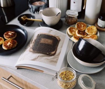High angle of cookbook and cooked fritters on plates placed on table near utensils and ingredients in cozy kitchen at home